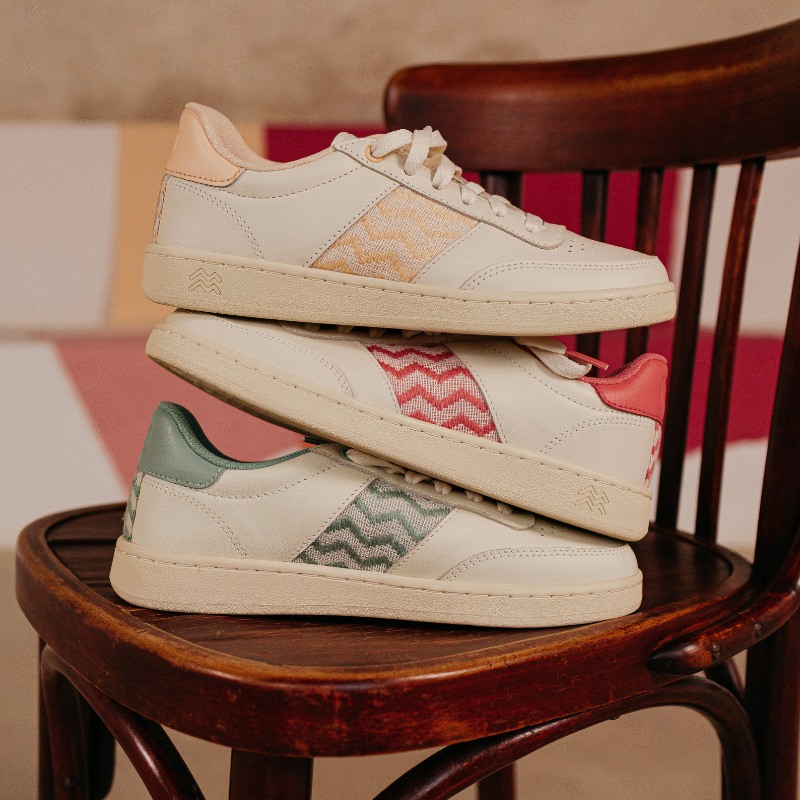 Three pairs of eco-friendly beige leather sneakers, stacked on a vintage wooden chair. Each shoe features a handwoven fabric detail with a zigzag pattern in pink, green, and beige. These sustainable sneakers combine minimalist style with natural materials