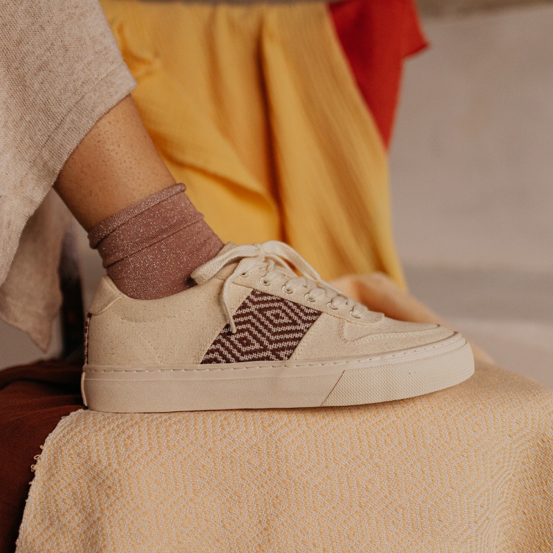 Close-up of a person wearing vegan beige leather sneakers, featuring a brown woven fabric pattern on the side. The shoe is paired with shimmering socks and an outfit in earthy tones. A pair of ethical sneakers that blend craftsmanship, comfort, and elegan